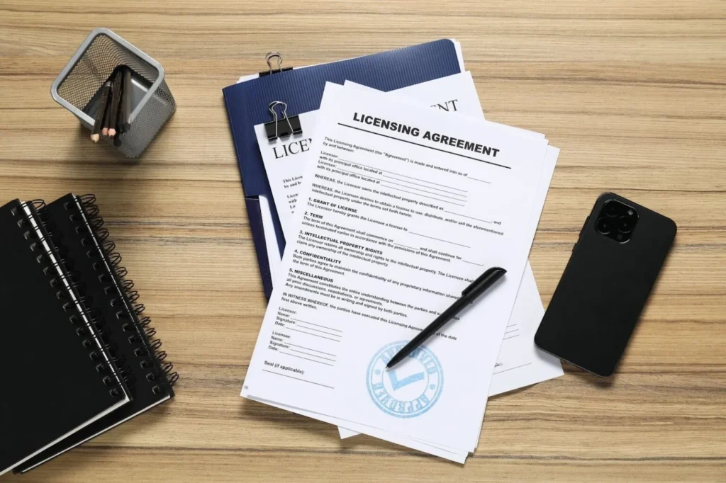 A licensing agreement document with an "Approved" stamp sitting on a wooden desk next to a smartphone, black notebook, and pen.