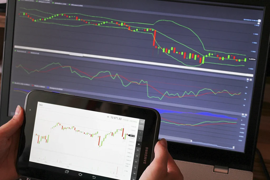 A person holding a Samsung tablet displaying a live candlestick stock chart next to a laptop screen showing technical analysis indicators, Bollinger Bands, and market trends for cryptocurrency trading.