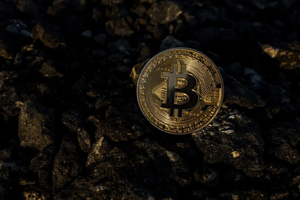 A physical gold Bitcoin coin resting on dark, jagged coal or volcanic rock, illuminated by a high-contrast side light to emphasize the "digital gold" and mining concept.