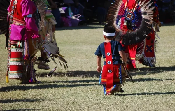 A young Indigenous boy in traditional regalia follows behind adult dancers in vibrant red and pink attire at an outdoor gathering, symbolizing the continuity of cultural wisdom.