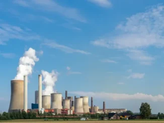 A wide landscape view of a large coal or gas power plant featuring multiple hyperbolic cooling towers and tall chimneys emitting thick white plumes of water vapor and smoke against a blue sky.