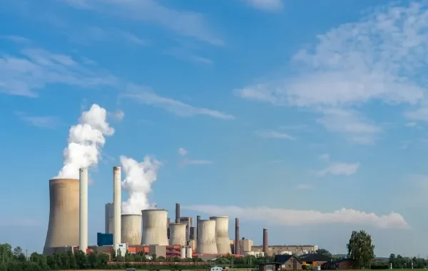 A wide landscape view of a large coal or gas power plant featuring multiple hyperbolic cooling towers and tall chimneys emitting thick white plumes of water vapor and smoke against a blue sky.
