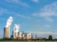 A wide landscape view of a large coal or gas power plant featuring multiple hyperbolic cooling towers and tall chimneys emitting thick white plumes of water vapor and smoke against a blue sky.