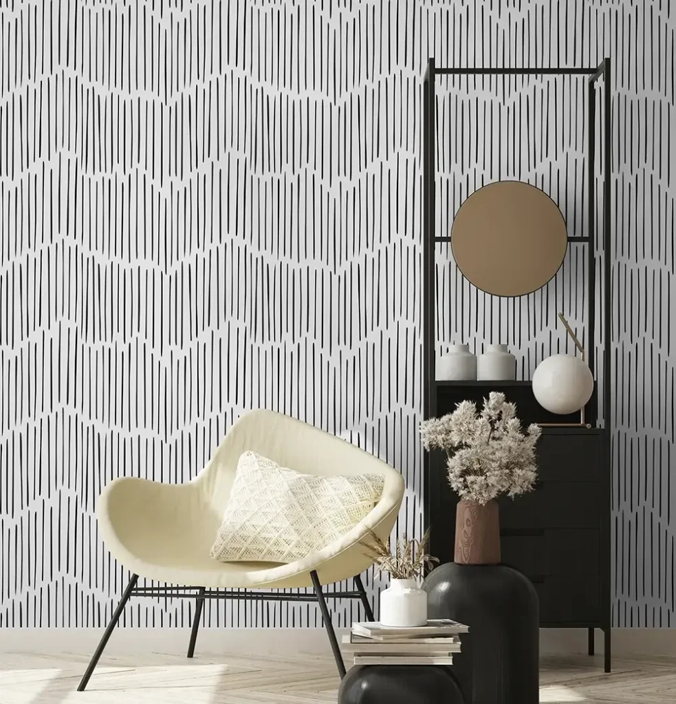 A stylish living room featuring modern black and white abstract vertical line wallpaper. The scene includes a cream-colored accent chair with a textured pillow, a minimalist black shelving unit with a round gold mirror, and neutral-toned decorative vases on a light herringbone wood floor.