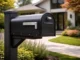A sleek, modern black metal mailbox mounted on a matching post in front of a contemporary home with a manicured green lawn and garden.