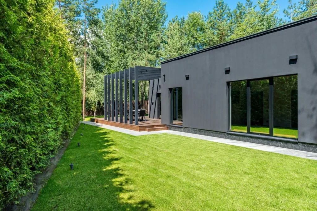 A contemporary, minimalist black garden room with large floor-to-ceiling windows, featuring a connected timber deck and a stylish dark wood pergola, set in a lush green backyard with a manicured lawn and tall hedges.