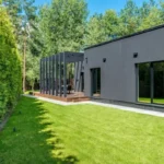 A contemporary dark charcoal garden room with large glass windows and a timber deck featuring a black pergola, set against a lush green lawn and high privacy hedge.
