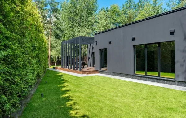 A contemporary dark charcoal garden room with large glass windows and a timber deck featuring a black pergola, set against a lush green lawn and high privacy hedge.