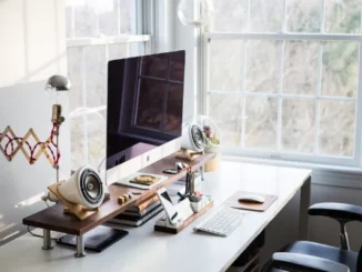 A bright, minimalist home office featuring an iMac on a wooden monitor riser, white cone-shaped speakers, a mechanical desk organizer, and a white desk positioned in front of a large window with natural light.