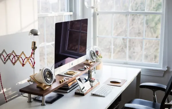A bright, minimalist home office featuring an iMac on a wooden monitor riser, white cone-shaped speakers, a mechanical desk organizer, and a white desk positioned in front of a large window with natural light.