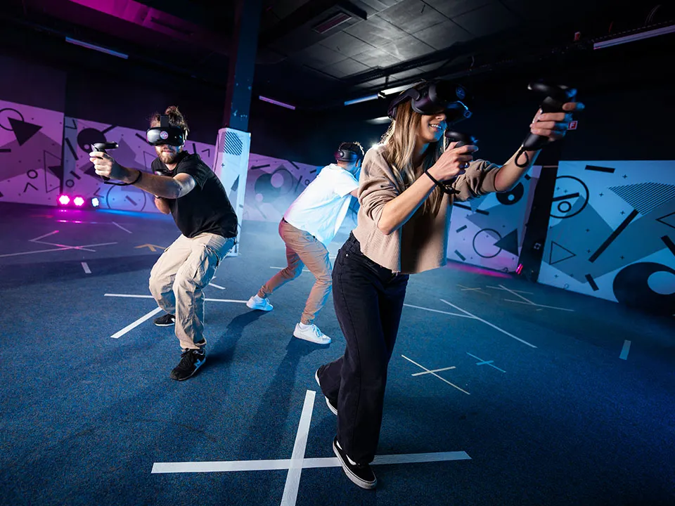 Three young adults wearing VR headsets and holding motion controllers actively play a team-based game in a large, professionally equipped virtual reality arena with neon lighting and floor tracking markers.