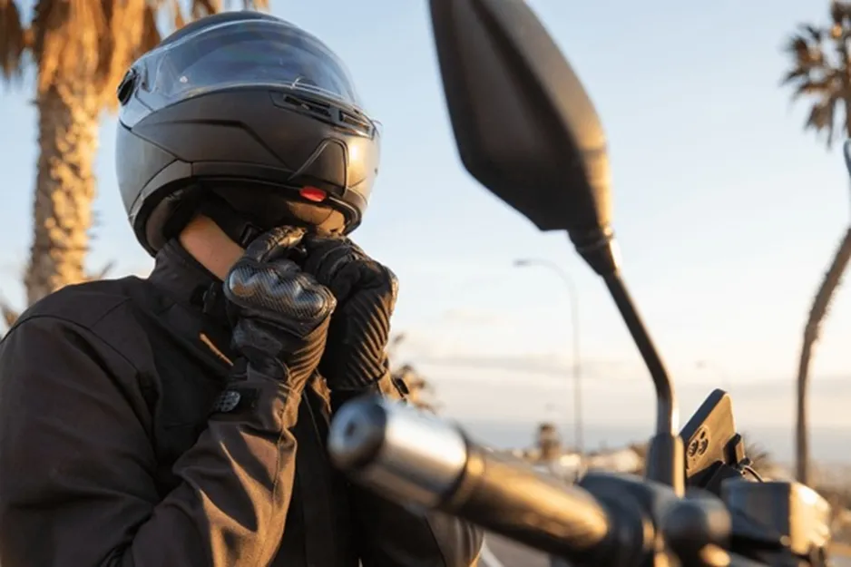 A motorcyclist in a black jacket and carbon fiber gloves fastening the chin strap of a matte black full-face helmet while seated on a bike.