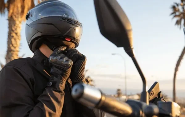 A motorcyclist in a black jacket and carbon fiber gloves fastening the chin strap of a matte black full-face helmet.