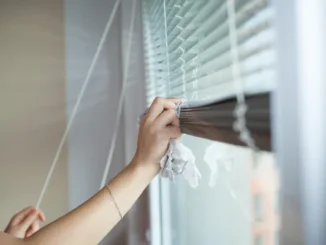 A person's hand using a microfiber cloth to carefully wipe dust from individual slats of horizontal window blinds.