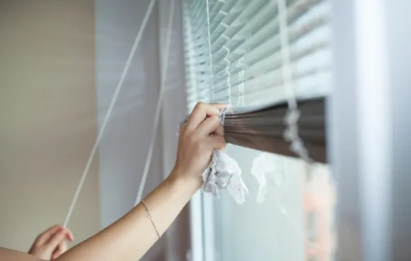 A person's hand using a microfiber cloth to carefully wipe dust from individual slats of horizontal window blinds.