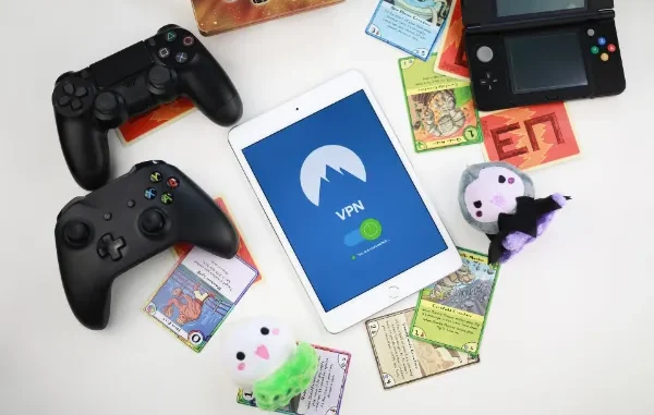 A top-down view of a white tablet displaying a VPN connection, surrounded by a PS4 controller, Xbox controller, Nintendo 3DS, trading cards, and plush toys.