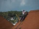 A professional contractor from a roofing company in Austin wearing a safety harness while inspecting a terracotta tile roof on a clear day.