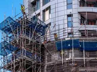 Professional siding contractors in safety gear working on a complex scaffolding system against a modern gray high-rise building.