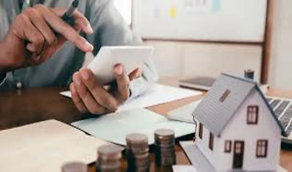 A person using a calculator at a desk with a small house model, coins, and documents, representing financial planning for property investments.