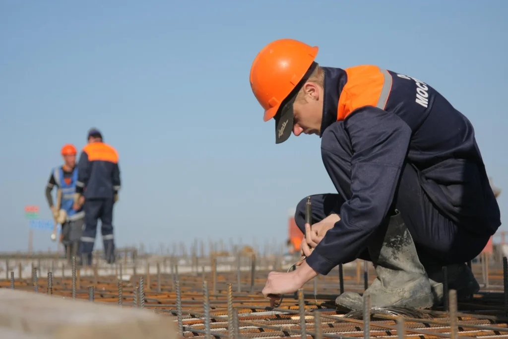 A professional siding contractor in a hard hat carefully inspecting and working on the structural foundation and exterior reinforcements of a residential building site.