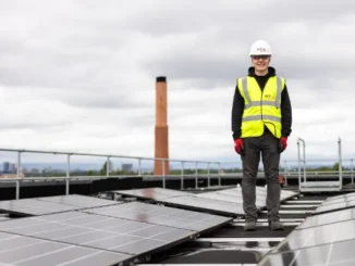 A solar technician in safety gear standing on a rooftop beside a large array of solar panels to ensure maximum energy efficiency.