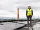 A solar technician in safety gear standing on a rooftop beside a large array of solar panels to ensure maximum energy efficiency.