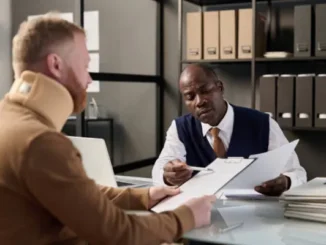 A personal injury lawyer in a vest and tie points to a document while consulting with a male client wearing a neck brace in an office.