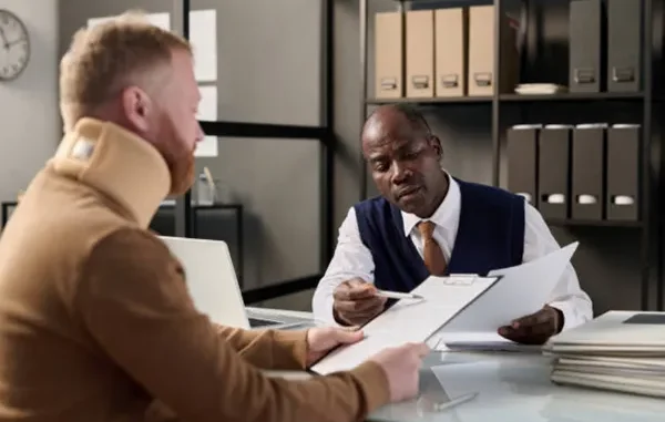 A personal injury lawyer in a vest and tie points to a document while consulting with a male client wearing a neck brace in an office.