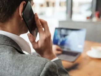 A businessman in a grey suit talking on a smartphone while monitoring data on a laptop in an office setting.