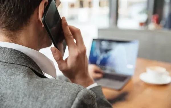 A businessman in a grey suit talking on a smartphone while monitoring data on a laptop in an office setting.