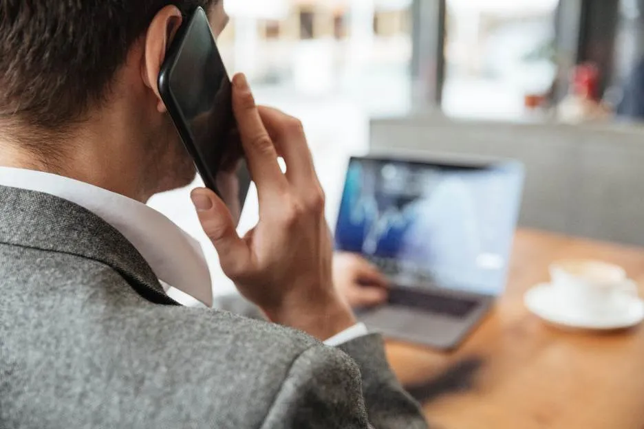 A businessman in a grey suit making a call on his smartphone while working on a laptop in a cafe, demonstrating the flexibility of T-Mobile call forwarding for remote work.