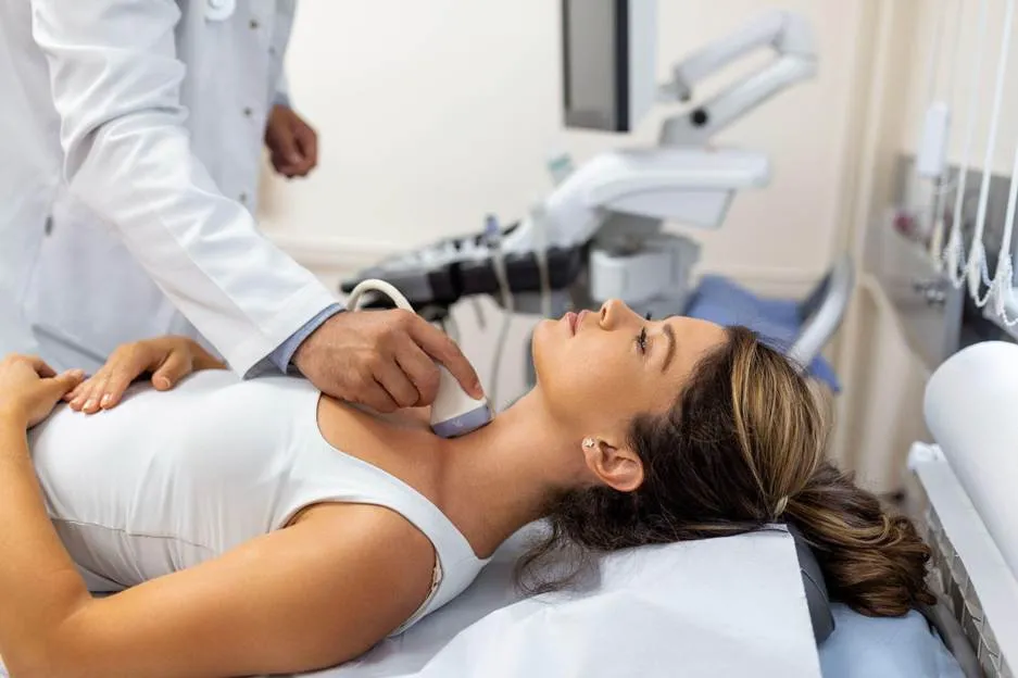 A medical professional performing an ultrasound on a woman's neck using a handheld transducer in a clinical setting.