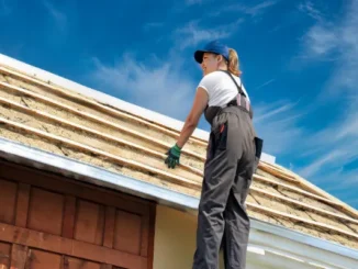 A female roofer in work gear standing on a ladder, inspecting the wooden structure and insulation of a roof under a clear blue sky.