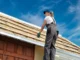 A female roofer in work gear standing on a ladder, inspecting the wooden structure and insulation of a roof under a clear blue sky.