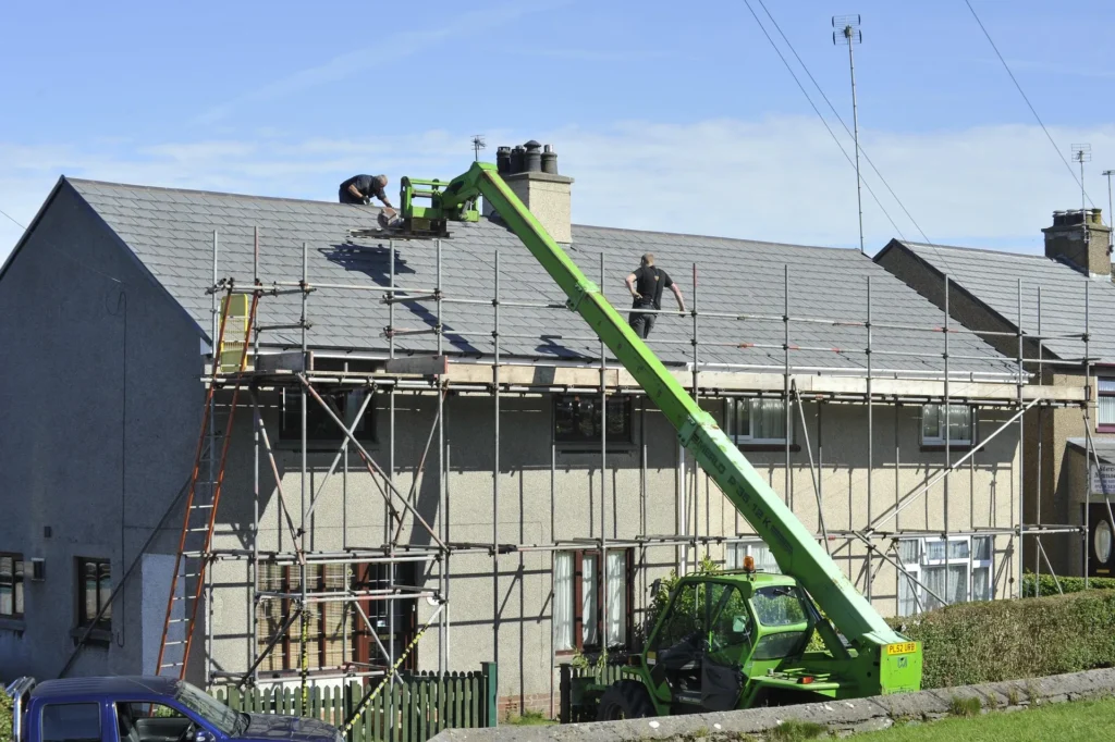 Two roofing contractors perform repairs on a grey slate roof of a residential house using extensive metal scaffolding and a green telescopic handler (telehandler) to lift materials.