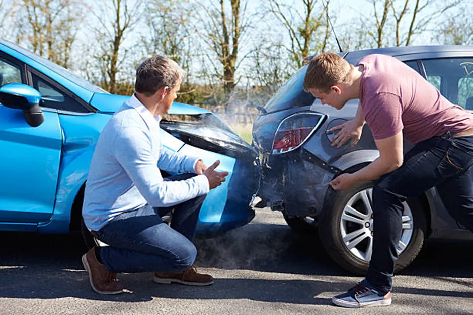 Two men inspecting a rear-end collision between a blue car and a grey car on the side of a road, showing visible bumper damage and smoke.