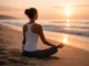 A woman sitting in a cross-legged yoga pose on a sandy beach, facing a glowing sunset over the ocean.