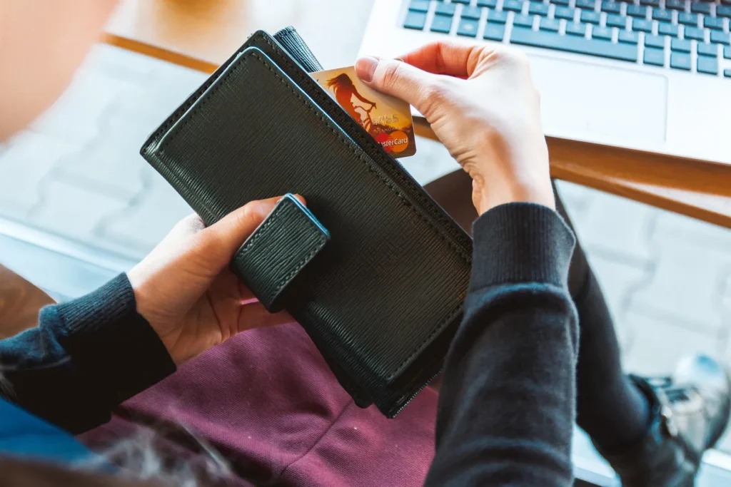 A person pulling a credit card out of a black wallet while sitting in front of a laptop, illustrating a secure online checkout process.