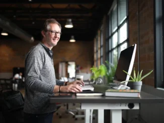 A smiling man working at a height-adjustable standing desk in a modern, open-concept office with large windows.