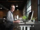 A smiling man working at a height-adjustable standing desk in a modern, open-concept office with large windows.