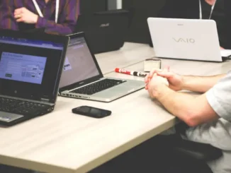 A group of startup founders and investors in a meeting with multiple laptops and a smartphone on a light wooden table, focusing on digital data analysis.