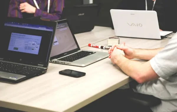 A group of startup founders and investors in a meeting with multiple laptops and a smartphone on a light wooden table, focusing on digital data analysis.