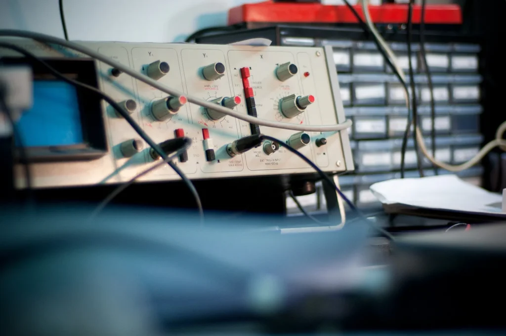 A close-up view of a vintage analog oscilloscope with various control knobs, switches, and BNC cables connected in a professional testing environment.