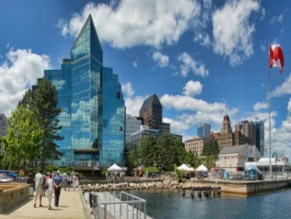 A sunny panoramic view of the Halifax waterfront in Nova Scotia, Canada, featuring the modern glass architecture of Queen’s Marque, a Canadian flag, and people walking along the boardwalk near the harbor.