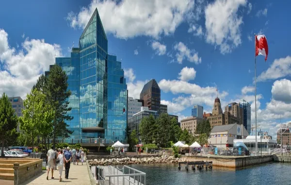 A sunny panoramic view of the Halifax waterfront in Nova Scotia, Canada, featuring the modern glass architecture of Queen’s Marque, a Canadian flag, and people walking along the boardwalk near the harbor.