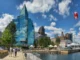 A sunny panoramic view of the Halifax waterfront in Nova Scotia, Canada, featuring the modern glass architecture of Queen’s Marque, a Canadian flag, and people walking along the boardwalk near the harbor.