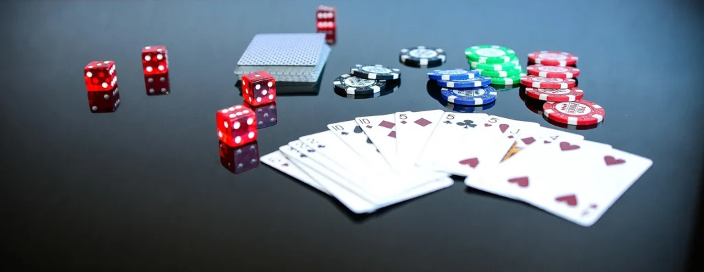 A professional casino table setup featuring a spread of poker cards, colorful betting chips, and red dice on a reflective dark surface.