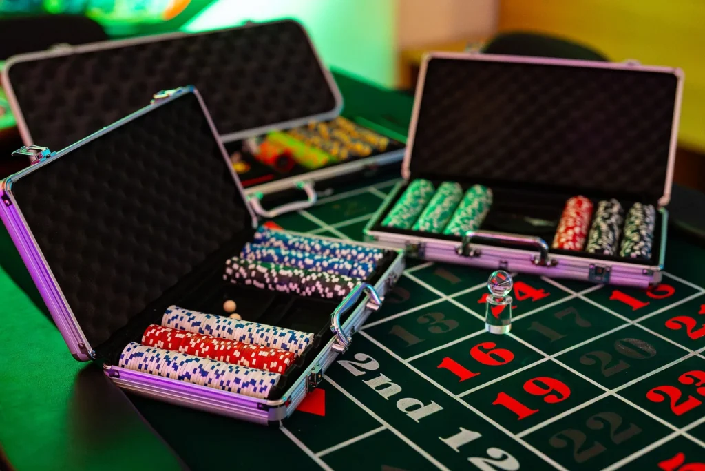Three open silver briefcases filled with colorful poker chips sitting on a green roulette table layout with numbered betting grids.