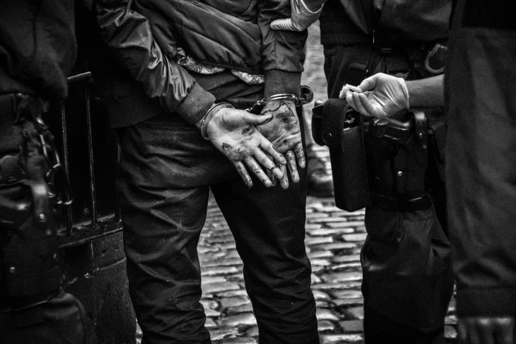 A black and white, close-up shot of a person's hands handcuffed behind their back. Their hands appear stained or dirty, and they are surrounded by law enforcement officers on a cobblestone street.