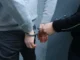A close-up shot showing a person’s hands being placed in metal handcuffs behind their back by another individual.